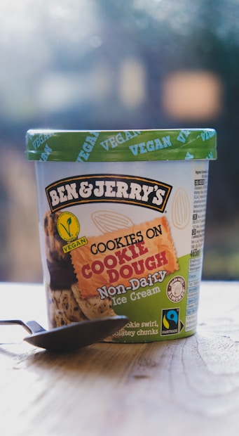 A container of Ben & Jerry's Cookies on Cookie Dough Non-Dairy vegan ice cream placed on a wooden surface. The container features a green lid with the word 'VEGAN' printed repeatedly. The label shows images of cookies and chocolate chunks and includes Fairtrade and non-dairy certifications. A spoon is placed in front of the container.
