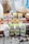 A selection of Foodie Flavours natural flavoring boxes are displayed on a wooden surface. Various flavors such as Rose, Apple, Lime, Pistachio, Organic Orange Oil, Apricot, Lemon, and Pineapple are showcased. In the background, there are baking ingredients like a measuring cup with brown sugar, a large mixing bowl with dough, a jug of milk, and colorful cupcake liners.