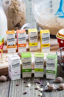 A selection of Foodie Flavours natural flavoring boxes are displayed on a wooden surface. Various flavors such as Rose, Apple, Lime, Pistachio, Organic Orange Oil, Apricot, Lemon, and Pineapple are showcased. In the background, there are baking ingredients like a measuring cup with brown sugar, a large mixing bowl with dough, a jug of milk, and colorful cupcake liners.