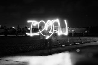 Light painting in a dark outdoor setting spells out 'I LOVE U' with bright white light against a black and white backdrop. The scene is nighttime with blurred greenery and structures in the background. There's a light trail effect creating a glowing text appearance on what seems to be a sports field.