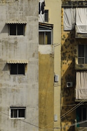 Old apartment building exterior with weathered and stained walls. The left side is pale gray with two small windows covered by awnings. The middle section is a faded yellow with clothes hanging near a small window and an air conditioning unit below. The right section has whitish sheets or curtains covering windows, with more stains becoming visible under sunlight.