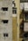 Old apartment building exterior with weathered and stained walls. The left side is pale gray with two small windows covered by awnings. The middle section is a faded yellow with clothes hanging near a small window and an air conditioning unit below. The right section has whitish sheets or curtains covering windows, with more stains becoming visible under sunlight.