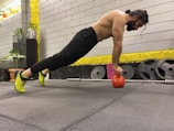 A man is performing an exercise in a gym, balancing his body in a plank position with his hands on a red kettlebell. He has a focused expression, suggesting concentration on his workout. The gym environment includes weight plates, a barbell, and potted plants for decoration.