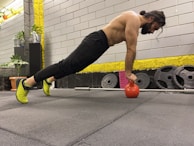 A man is performing an exercise in a gym, balancing his body in a plank position with his hands on a red kettlebell. He has a focused expression, suggesting concentration on his workout. The gym environment includes weight plates, a barbell, and potted plants for decoration.