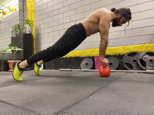 A man is performing an exercise in a gym, balancing his body in a plank position with his hands on a red kettlebell. He has a focused expression, suggesting concentration on his workout. The gym environment includes weight plates, a barbell, and potted plants for decoration.