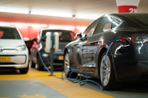 Several electric cars are charging in an indoor parking garage. The space is brightly lit with predominant red and white colors in the background. Charging cables are connected to the cars, highlighting a modern and technological atmosphere.