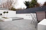 A modern outdoor patio area with grey tiled flooring and a minimalist design. The space features two sleek metal chairs and white planters with small trees and flowers. A black horizontal-slat fence adds privacy, while trees and shrubs provide greenery in the background.