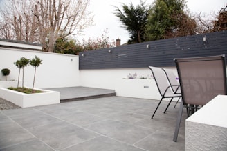 A modern outdoor patio area with grey tiled flooring and a minimalist design. The space features two sleek metal chairs and white planters with small trees and flowers. A black horizontal-slat fence adds privacy, while trees and shrubs provide greenery in the background.