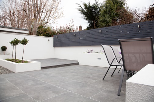 A modern outdoor patio area with grey tiled flooring and a minimalist design. The space features two sleek metal chairs and white planters with small trees and flowers. A black horizontal-slat fence adds privacy, while trees and shrubs provide greenery in the background.