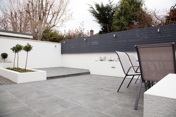 A modern outdoor patio area with grey tiled flooring and a minimalist design. The space features two sleek metal chairs and white planters with small trees and flowers. A black horizontal-slat fence adds privacy, while trees and shrubs provide greenery in the background.