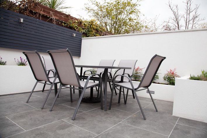 A modern outdoor patio setting features a rectangular glass-top table surrounded by six sleek metal chairs with mesh backs and seats. The patio is paved with large gray tiles, and the area is enclosed by white walls with some greenery peeking at the top, along with some metal detailing for water drainage.