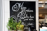 A chalkboard with elegant cursive writing in German is on display, surrounded by white tiles. In front of the chalkboard, there are several pots of green plants. On the right, shelves hold stacks of white cups and bowls, and a spotlight fixture is mounted on the ceiling.