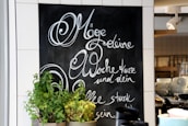 A chalkboard with elegant cursive writing in German is on display, surrounded by white tiles. In front of the chalkboard, there are several pots of green plants. On the right, shelves hold stacks of white cups and bowls, and a spotlight fixture is mounted on the ceiling.
