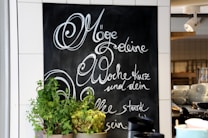 A chalkboard with elegant cursive writing in German is on display, surrounded by white tiles. In front of the chalkboard, there are several pots of green plants. On the right, shelves hold stacks of white cups and bowls, and a spotlight fixture is mounted on the ceiling.