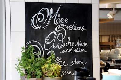 A chalkboard with elegant cursive writing in German is on display, surrounded by white tiles. In front of the chalkboard, there are several pots of green plants. On the right, shelves hold stacks of white cups and bowls, and a spotlight fixture is mounted on the ceiling.