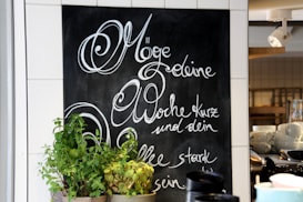 A chalkboard with elegant cursive writing in German is on display, surrounded by white tiles. In front of the chalkboard, there are several pots of green plants. On the right, shelves hold stacks of white cups and bowls, and a spotlight fixture is mounted on the ceiling.