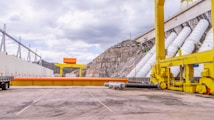A large industrial facility featuring a dam with multiple large white pipes descending from the structure. The foreground shows an expansive concrete area with yellow machinery, including a crane structure and a flatbed trailer. A mountainous terrain is visible in the background under a cloudy sky.