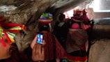 A group of people is walking through a narrow, rocky passage. They are wearing colorful, traditional clothing adorned with bright patterns and some are wearing hats decorated with feathers. One person in the foreground is holding a smartphone, capturing a photo of the scene.