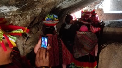 A group of people is walking through a narrow, rocky passage. They are wearing colorful, traditional clothing adorned with bright patterns and some are wearing hats decorated with feathers. One person in the foreground is holding a smartphone, capturing a photo of the scene.