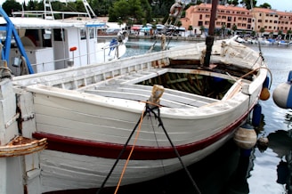 A wooden boat is docked at a harbor with a worn, rustic appearance. It is primarily white with some red accents and is secured with ropes. Nearby, a modern white boat is visible. The harbor is calm, and the scene includes various buildings and trees in the background.