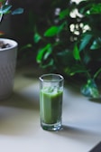 A tall glass filled with a green smoothie sits on a light-colored table. In the background, lush green foliage adds a natural and fresh atmosphere.