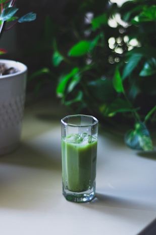 A tall glass filled with a green smoothie sits on a light-colored table. In the background, lush green foliage adds a natural and fresh atmosphere.