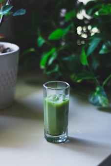A tall glass filled with a green smoothie sits on a light-colored table. In the background, lush green foliage adds a natural and fresh atmosphere.