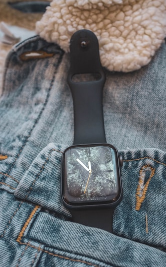 A black smartwatch with a worn, textured screen is resting on a pair of blue denim jeans. The jeans feature some visible stitching and a torn area, giving a rustic and casual feel. A beige, fuzzy fabric is partially visible in the background, adding a cozy texture to the composition.