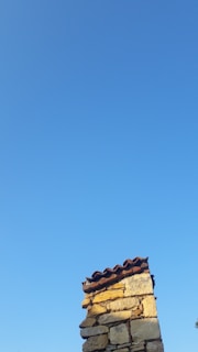 A clear blue sky with a rustic stone chimney extending upwards from the bottom of the frame. The chimney is made of rough-hewn stones and topped with a row of clay tiles. The sunlight casts warm, yellowish hues on the stones, contrasting with the vivid blue of the sky.