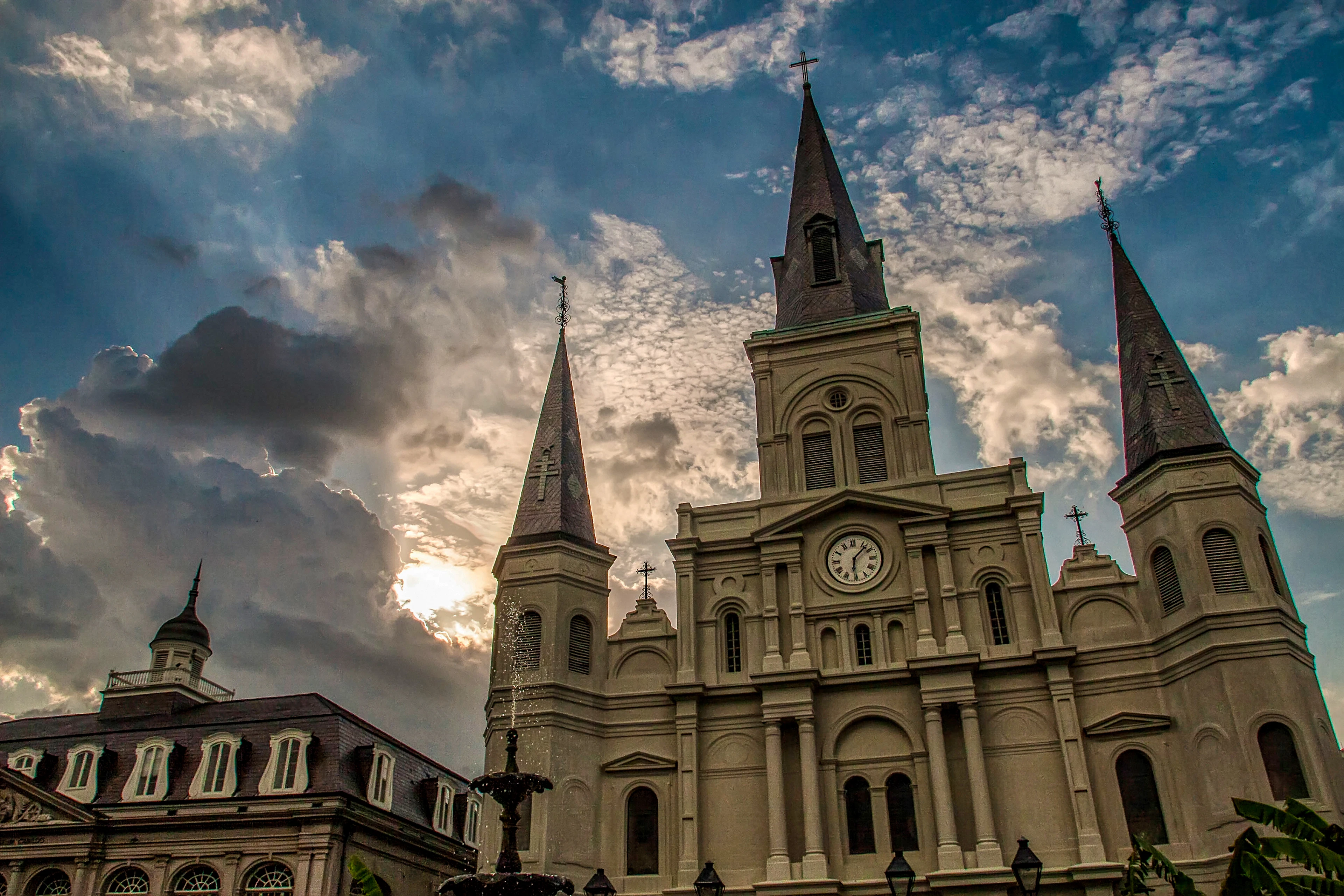 St. Louis Cathedral on Jackson Square in New Orleans, its three slate spires and clock face pale against a sky of towering clouds with the sun burning through behind them