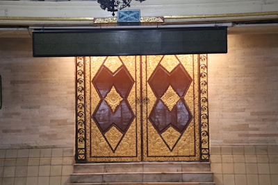 An ornate double door with intricate carvings and geometric patterns in gold and rich brown tones. The wall surrounding the door consists of light-colored brick or stone, complemented by a few steps leading up to the entrance.