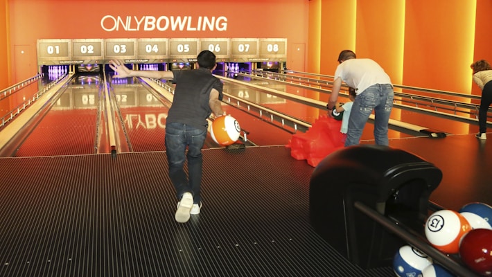 People are enjoying a game of bowling in a modern alley. The space is warmly lit with orange tones and features multiple lanes, each with an overhead screen displaying scores. Bowling balls are visible, including an eight ball being held by a player ready to roll. Another player is setting up his turn, and there are people in the background, focusing on their games.