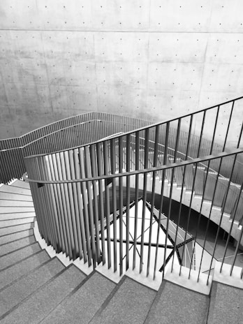 A modern architectural interior featuring a spiral staircase with sleek metal railings and concrete walls. The spiral design creates a sense of movement and complexity, enhanced by the shadows and light patterns on the surface.