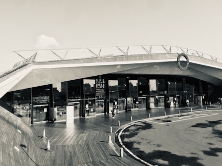 A modern architectural building with sleek, angular lines and large glass windows. The structure features a rooftop deck with railings. People can be seen walking near the entrance, and traffic cones are placed on a wooden deck area, adding an industrial yet clean aesthetic.