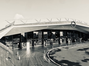 A modern architectural building with sleek, angular lines and large glass windows. The structure features a rooftop deck with railings. People can be seen walking near the entrance, and traffic cones are placed on a wooden deck area, adding an industrial yet clean aesthetic.