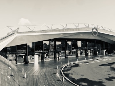 A modern architectural building with sleek, angular lines and large glass windows. The structure features a rooftop deck with railings. People can be seen walking near the entrance, and traffic cones are placed on a wooden deck area, adding an industrial yet clean aesthetic.