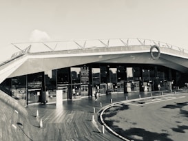 A modern architectural building with sleek, angular lines and large glass windows. The structure features a rooftop deck with railings. People can be seen walking near the entrance, and traffic cones are placed on a wooden deck area, adding an industrial yet clean aesthetic.