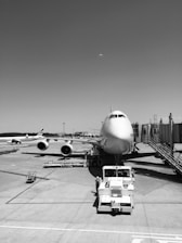 An airplane is parked at an airport gate, connected by a jet bridge. Ground service vehicles and other infrastructure are visible, including a tug vehicle directly in front of the plane. Several other planes can be seen in the background on the tarmac.