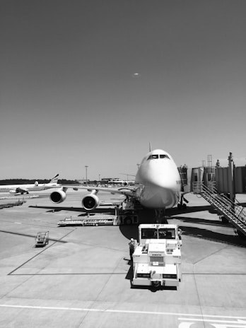 An airplane is parked at an airport gate, connected by a jet bridge. Ground service vehicles and other infrastructure are visible, including a tug vehicle directly in front of the plane. Several other planes can be seen in the background on the tarmac.