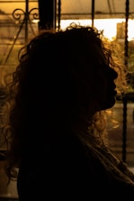 A silhouette of a person with curly hair against a warm, glowing sunset backdrop. The light filters through what appears to be a window with intricate ironwork.