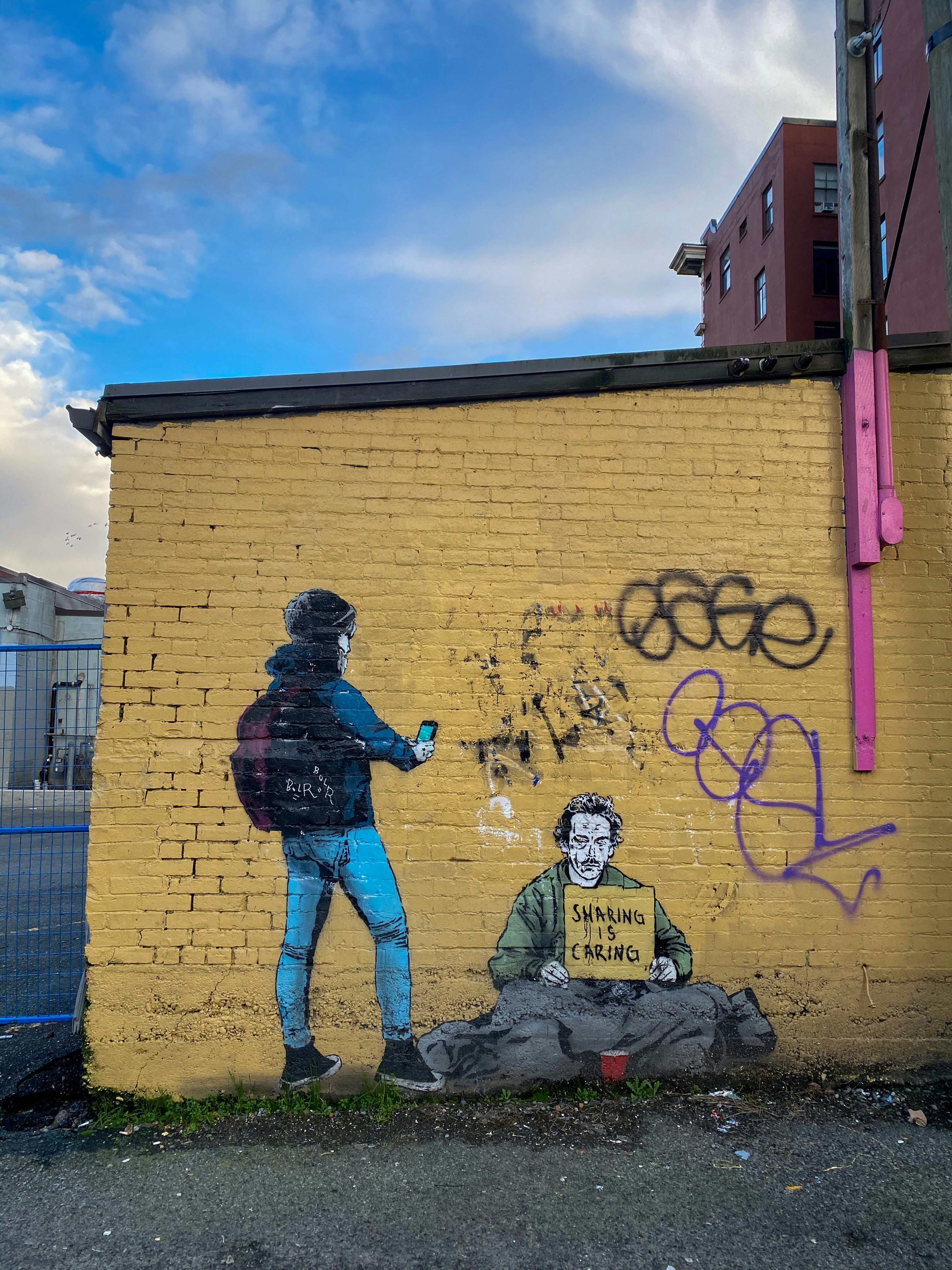A mural on a yellow brick wall depicts a person wearing a beanie and backpack holding a phone, standing beside a homeless man sitting on the ground. The homeless man holds a sign that reads 'Sharing is Caring'. The wall also features graffiti and has a clear, blue sky above it.