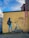 A mural on a yellow brick wall depicts a person wearing a beanie and backpack holding a phone, standing beside a homeless man sitting on the ground. The homeless man holds a sign that reads 'Sharing is Caring'. The wall also features graffiti and has a clear, blue sky above it.