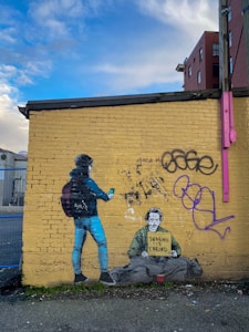 A mural on a yellow brick wall depicts a person wearing a beanie and backpack holding a phone, standing beside a homeless man sitting on the ground. The homeless man holds a sign that reads 'Sharing is Caring'. The wall also features graffiti and has a clear, blue sky above it.