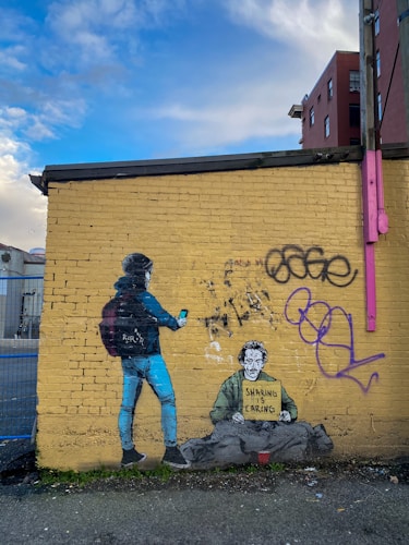 A mural on a yellow brick wall depicts a person wearing a beanie and backpack holding a phone, standing beside a homeless man sitting on the ground. The homeless man holds a sign that reads 'Sharing is Caring'. The wall also features graffiti and has a clear, blue sky above it.