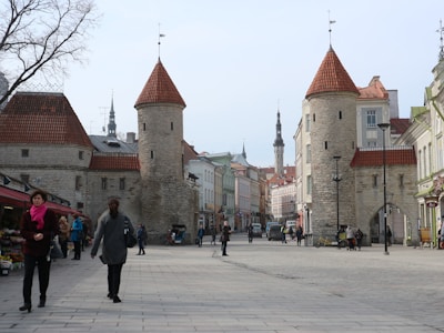 A street scene in a medieval European city with stone towers and red-tiled rooftops lining the street. Several people are walking and shopping, and there are colorful buildings in the background.