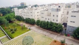 A residential area with several white apartment buildings surrounded by greenery. There is a well-maintained garden with trees and a paved walkway. The foreground features a geometric dome structure on a patterned surface. The background shows other city buildings.