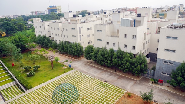 A residential area with several white apartment buildings surrounded by greenery. There is a well-maintained garden with trees and a paved walkway. The foreground features a geometric dome structure on a patterned surface. The background shows other city buildings.
