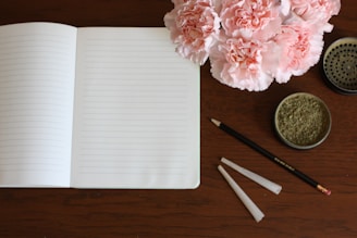 A wooden table surface features an open, lined notebook alongside a black pencil with an eraser. To the right, a container filled with ground herb is present, and above it, there is a round metal grinder. Adjacent to these items, two empty, rolled paper cones are laid out. A bouquet of pink carnations adds a touch of color to the scene.
