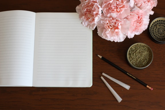 A wooden table surface features an open, lined notebook alongside a black pencil with an eraser. To the right, a container filled with ground herb is present, and above it, there is a round metal grinder. Adjacent to these items, two empty, rolled paper cones are laid out. A bouquet of pink carnations adds a touch of color to the scene.