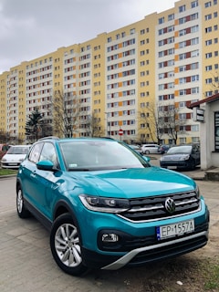 A teal Volkswagen SUV is parked on a concrete surface in front of a tall, yellow residential apartment building. The building has multiple floors with identical windows and balconies, and there are other vehicles parked nearby. Trees with sparse branches are visible in front of the building under a cloudy sky.